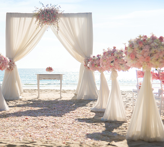 Beach wedding ceremony setup with a white draped arch decorated with pink and white flowers, facing the ocean with chairs on the sides.