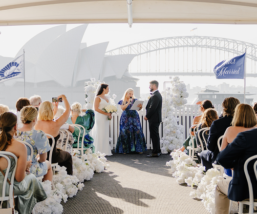 A wedding ceremony taking place outdoors with the Sydney Opera House and Harbour Bridge in the background. The bride and groom are exchanging vows, surrounded by seated guests, white flowers, and a 'Just Married' banner.