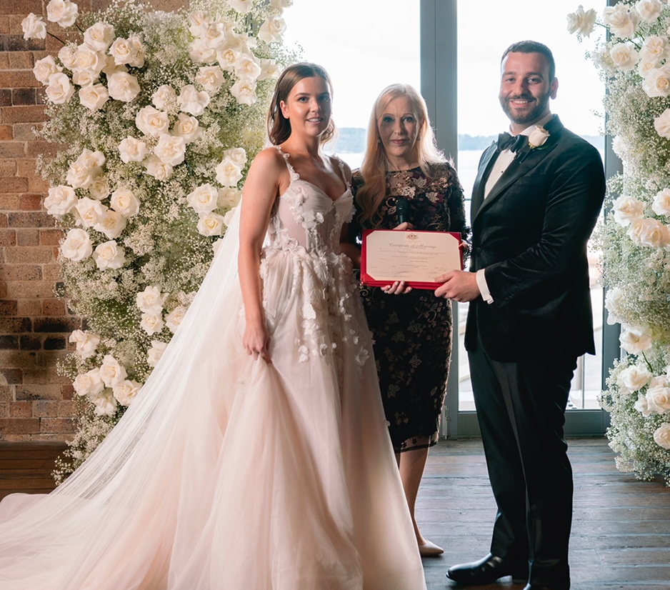 A bride and groom are standing with a woman, possibly an officiant, at a wedding ceremony. They are holding a certificate, and there are white floral arrangements and a window in the background.