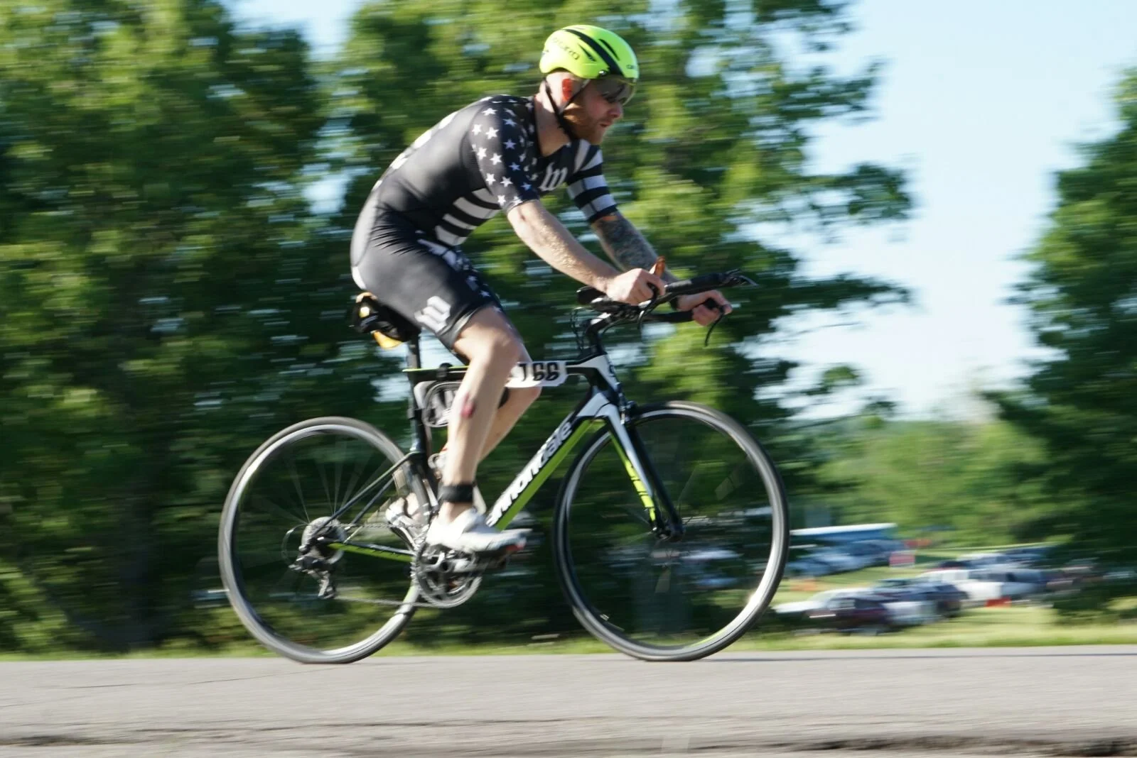 A man riding a road bicycle on a paved path, wearing a yellow helmet, sunglasses, and a black and white striped cycling jersey with stars, with trees and cars blurred in the background.