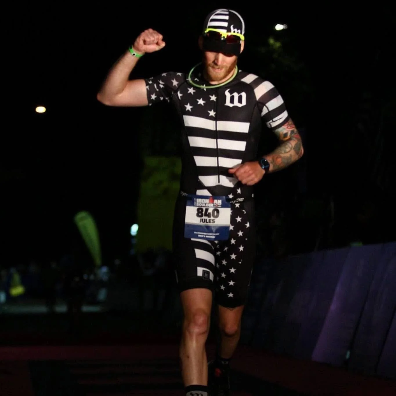 Man wearing a black and white American flag-themed sports outfit, sunglasses, and a race bib number 840, running at night with his right arm flexed.