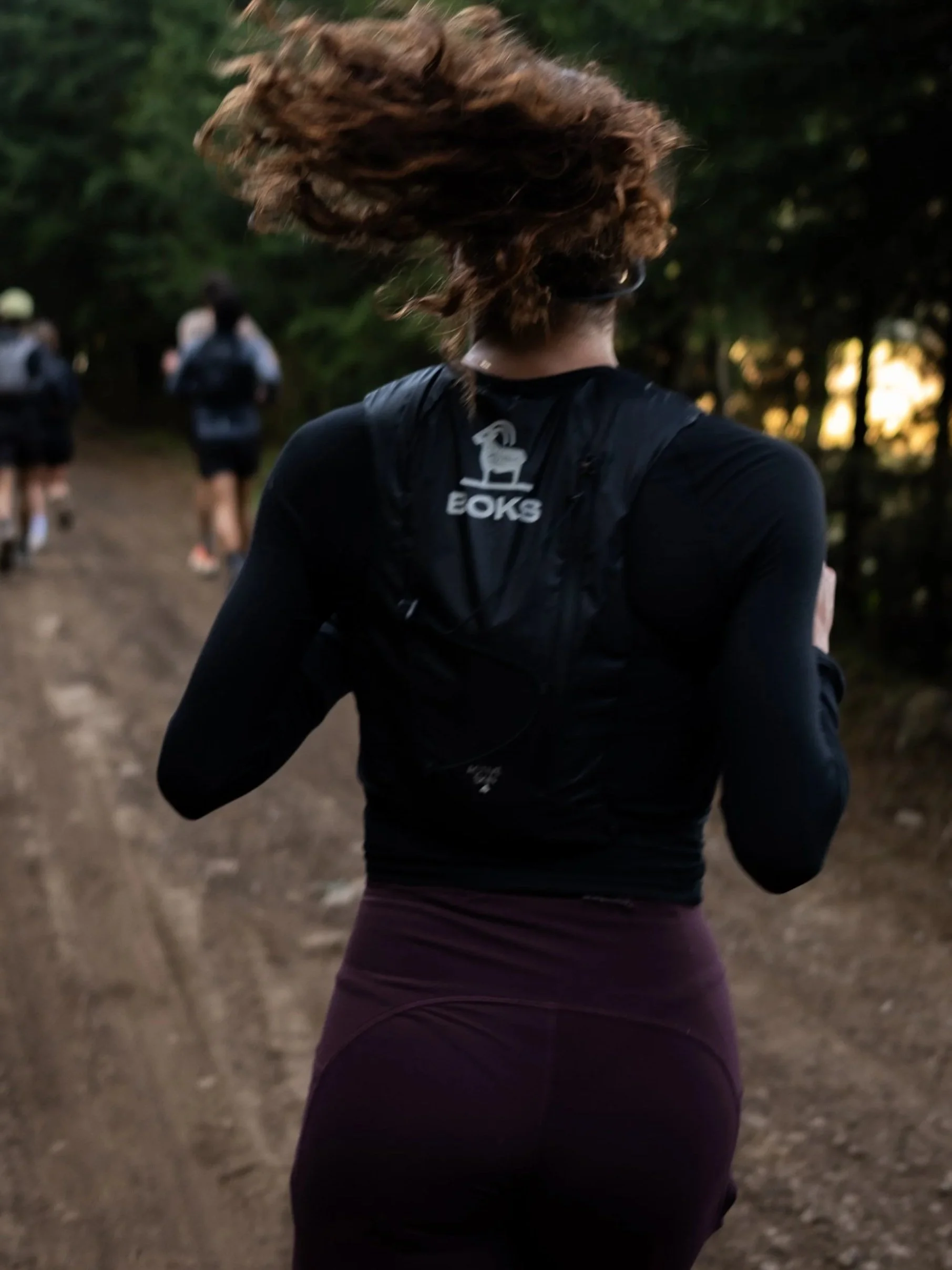 Mujer corriendo en un sendero en un bosque, vista desde atrás con logo de BOKS . trc en su chaleco.