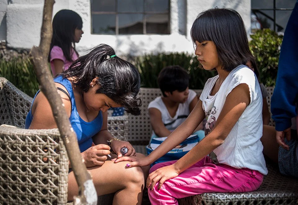 Una niña pintando las uñas de la mano derecha de otra niña. Otras niñas están sentadas en el fondo en un espacio al aire libre.