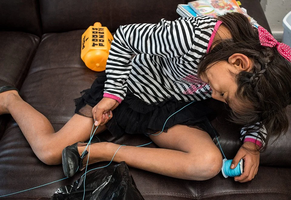 Una niña en una pose peculiar, acostada en un mueble de color marrón, sosteniendo un carrete de hilo azul, con varios libros y un balón de agua amarillo al fondo.