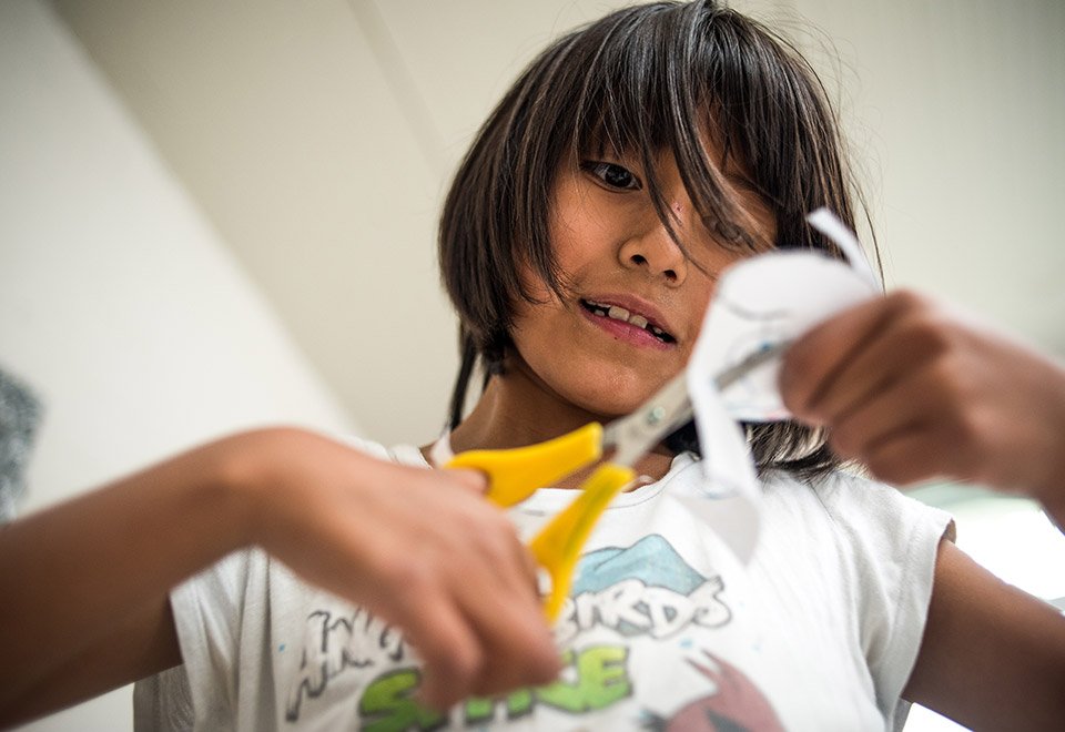 Niña con cabello castaño oscuro cortó y desordenado, usando tijeras para cortar un papel o cuerda, con expresión de concentración.