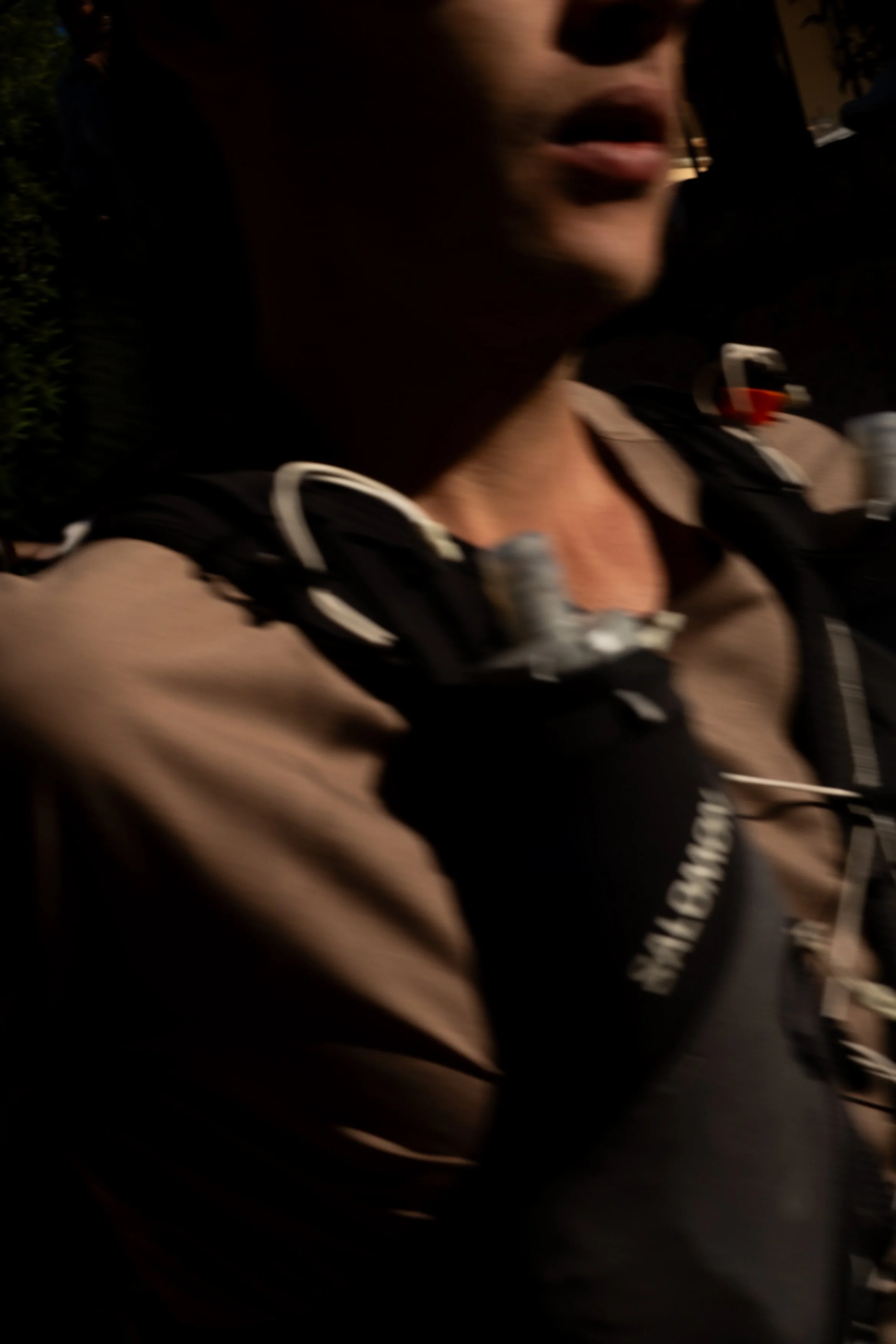 Close-up of a person's face, slightly blurred, wearing a brown jacket and a black backpack with white straps.
