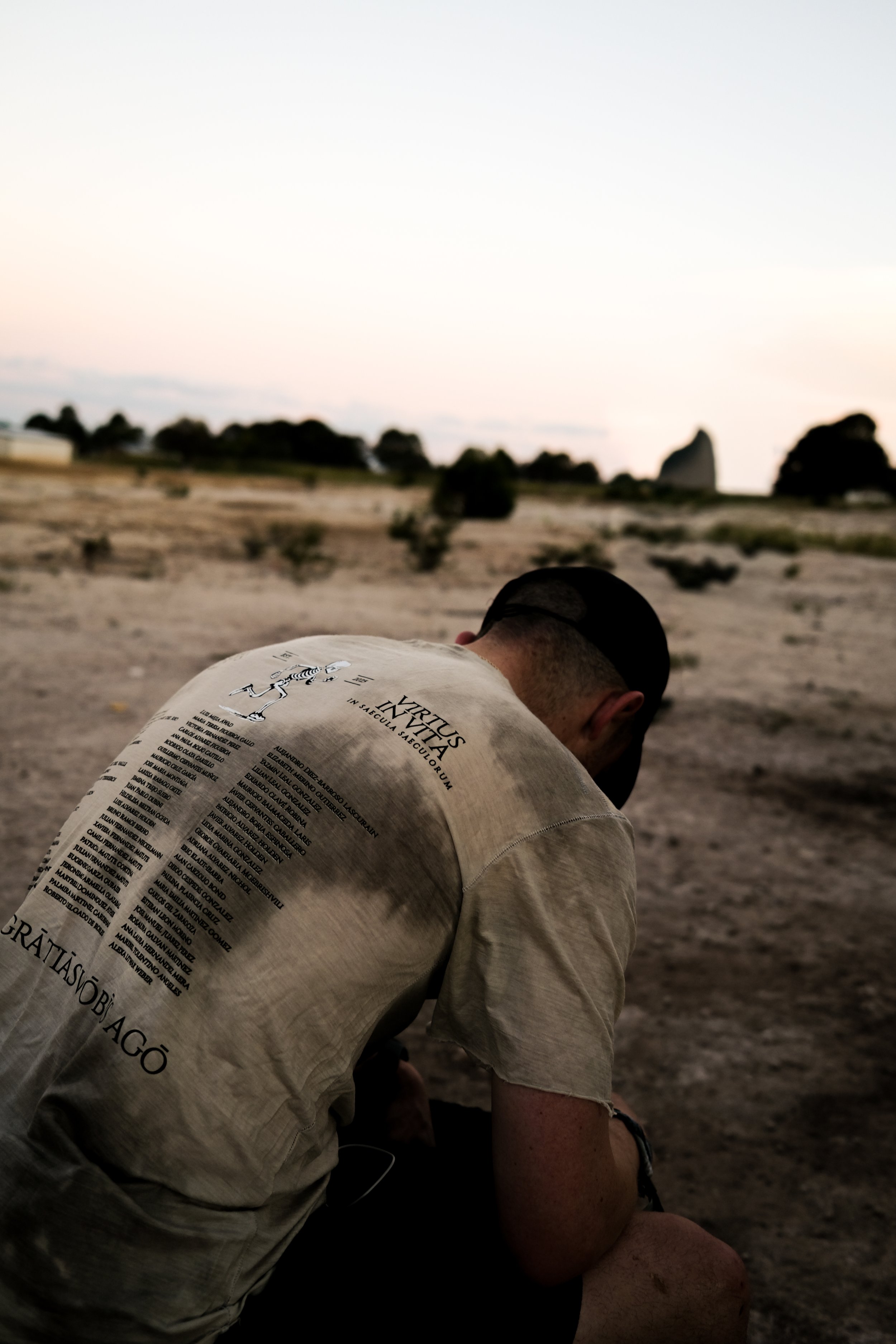A man with a beard and wearing a cap sits in a desert landscape during sunset, facing away from the camera.