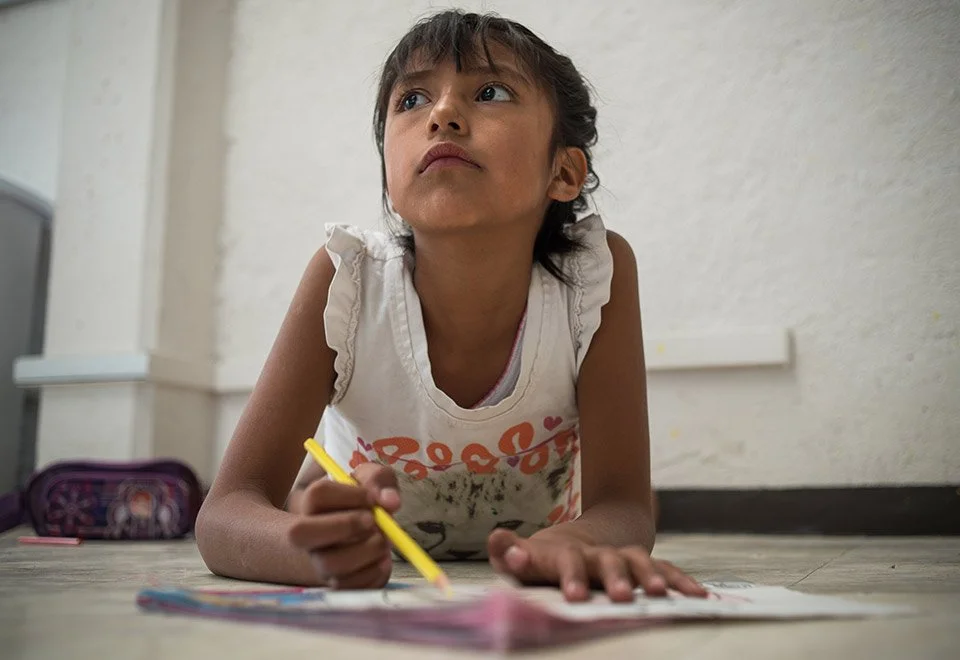 Niña acostada en el suelo, sosteniendo un lápiz amarillo y apuntando a un cuaderno o libro, parece estar pensando o concentrada.