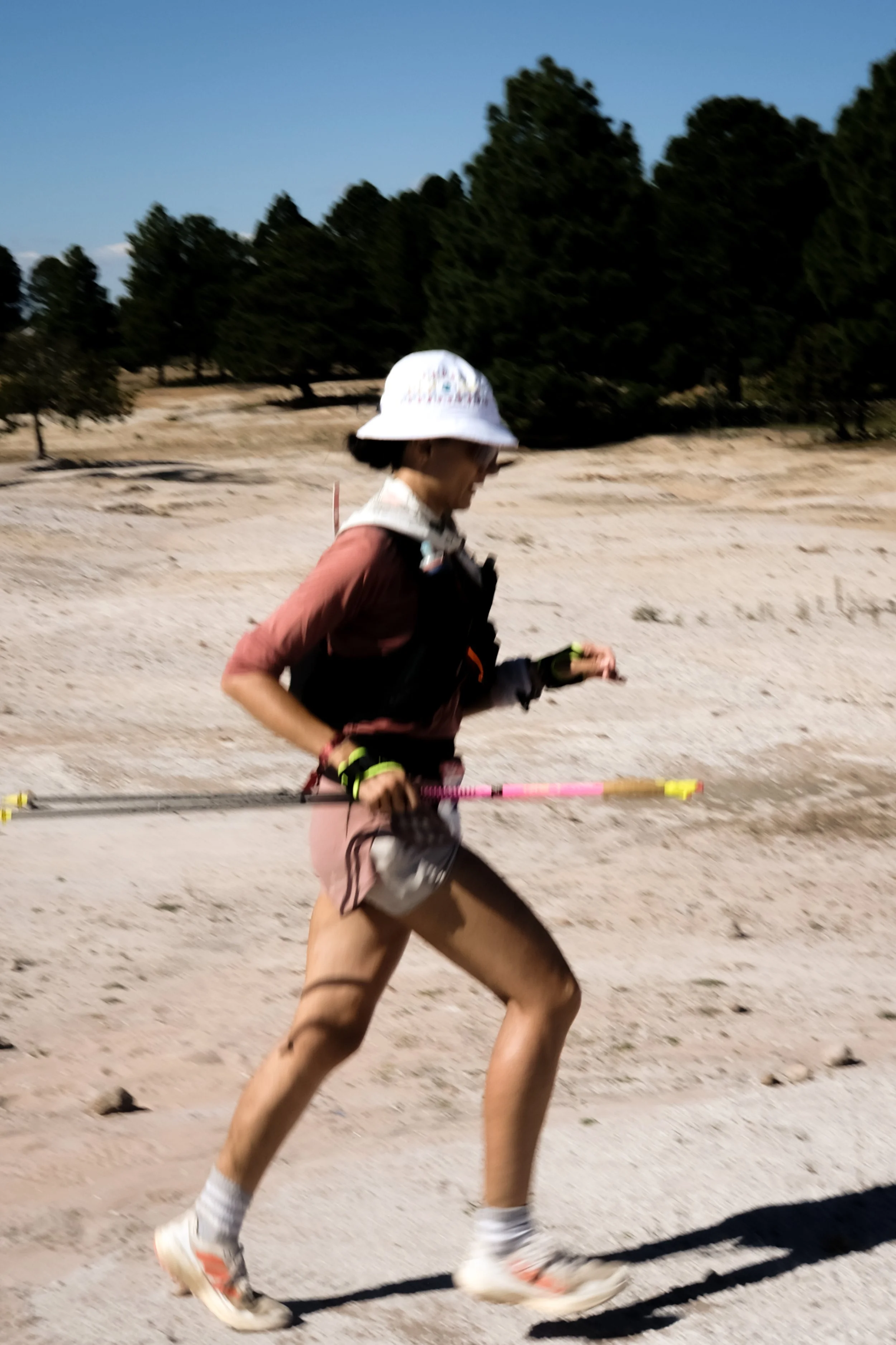 A woman running outdoors on a dirt trail with trees in the background, wearing a white hat, sunglasses, a long-sleeve shirt, shorts, and holding trekking poles.
