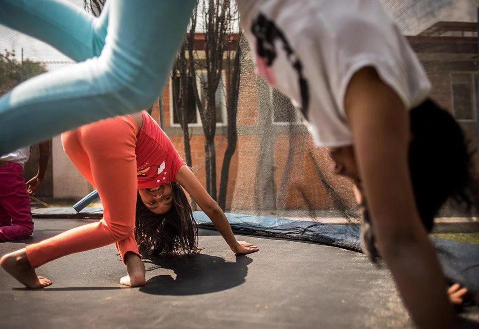 Niños jugando en una cama elástica, uno de ellos haciendo una voltereta hacia adelante, en un ambiente exterior con mallas de protección y árboles en el fondo.