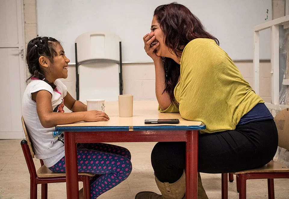 Una mujer y una niña riendo juntas en una mesa, compartiendo un momento alegre.
