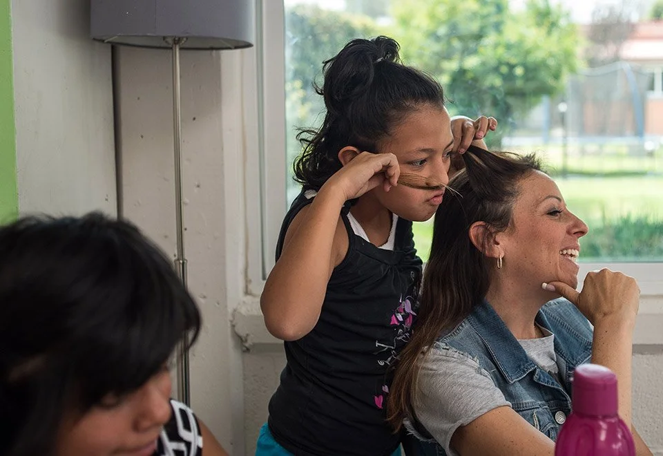 Una niña jugando con una mujer, en un espacio interior junto a una ventana. La niña tiene el cabello recogido y está haciendo una expresión divertida, mientras la mujer se ríe.