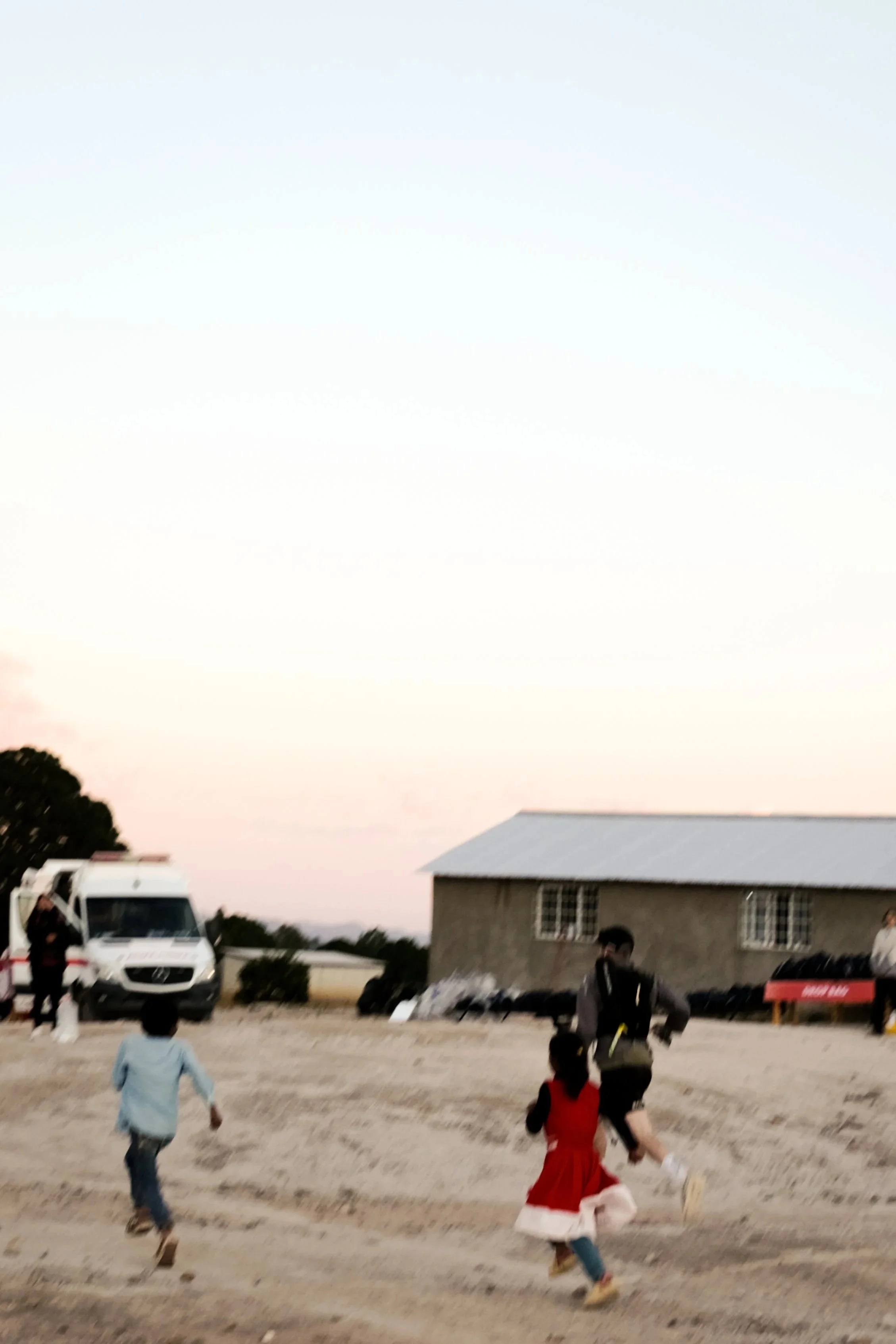 Niños corriendo en un campo exterior al atardecer, con una casa y un vehículo en el fondo.