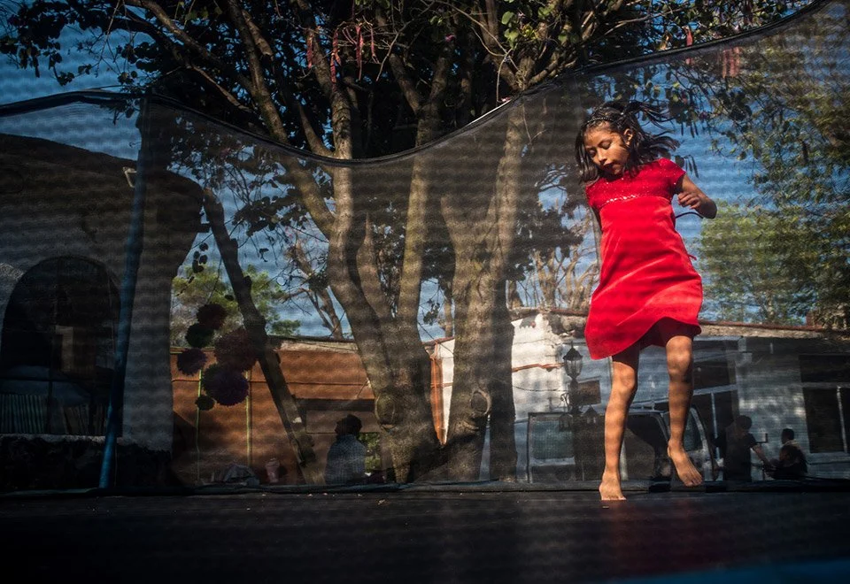 Una niña con vestido rojo brinca en una cama elástica en un patio con árboles y estructuras en el fondo, vista desde abajo a través del tapiz de la cama elástica.