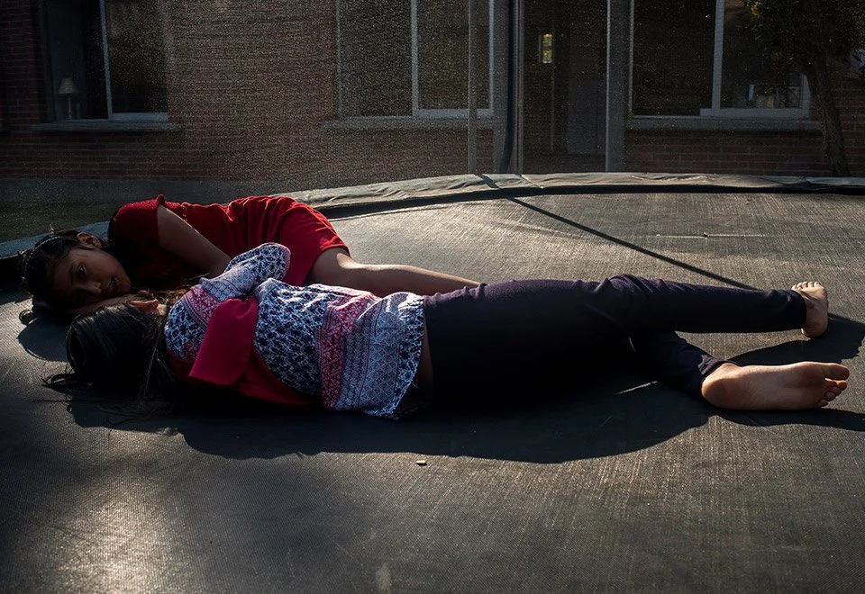 Dos niñas acostadas juntas en una cama elástica al aire libre, una mira hacia la cámara y la otra tiene los ojos cerrados.