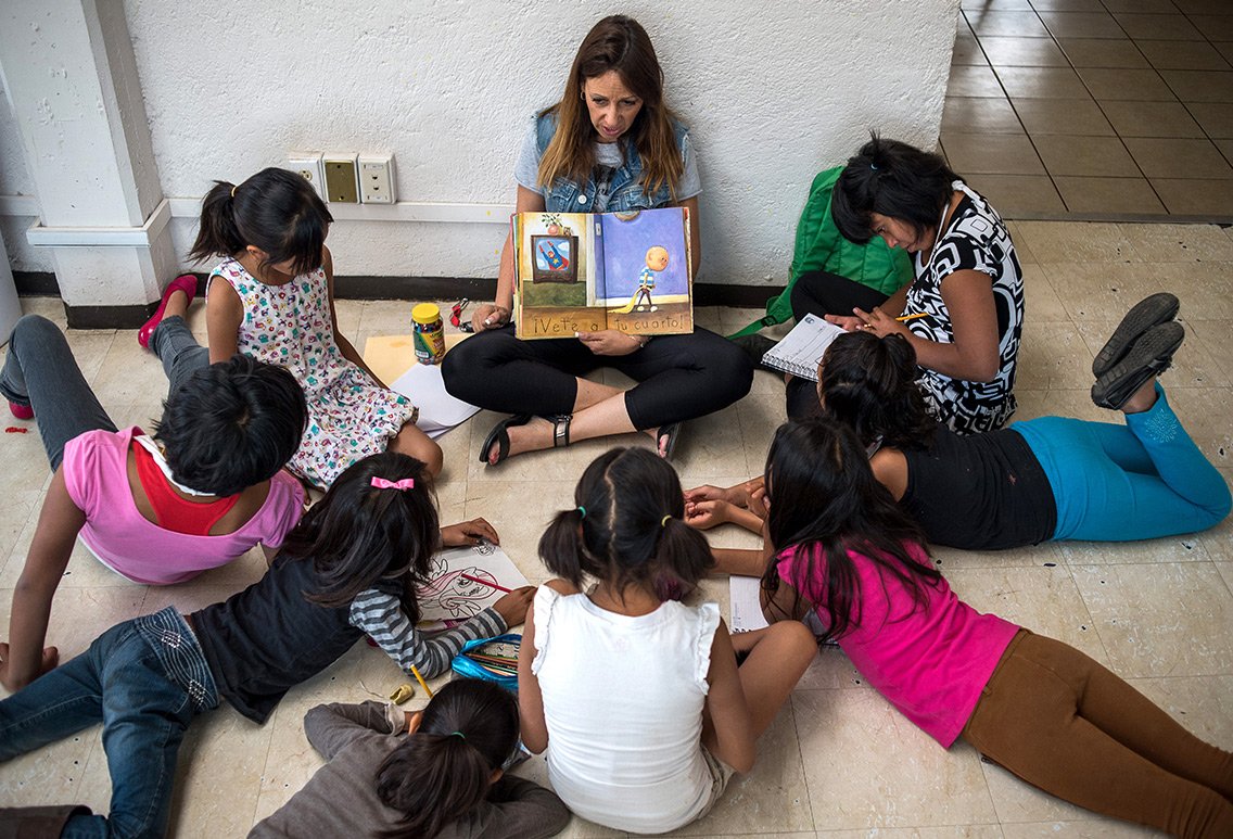 Una mujer lee un libro infantil a un grupo de niños que están acostados en el suelo, algunos dibujan y otros toman notas.