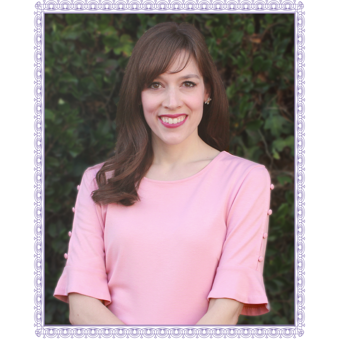 A woman with light skin and long brown hair smiling, wearing a pink top with ruffled sleeves, standing outdoors in front of green foliage.