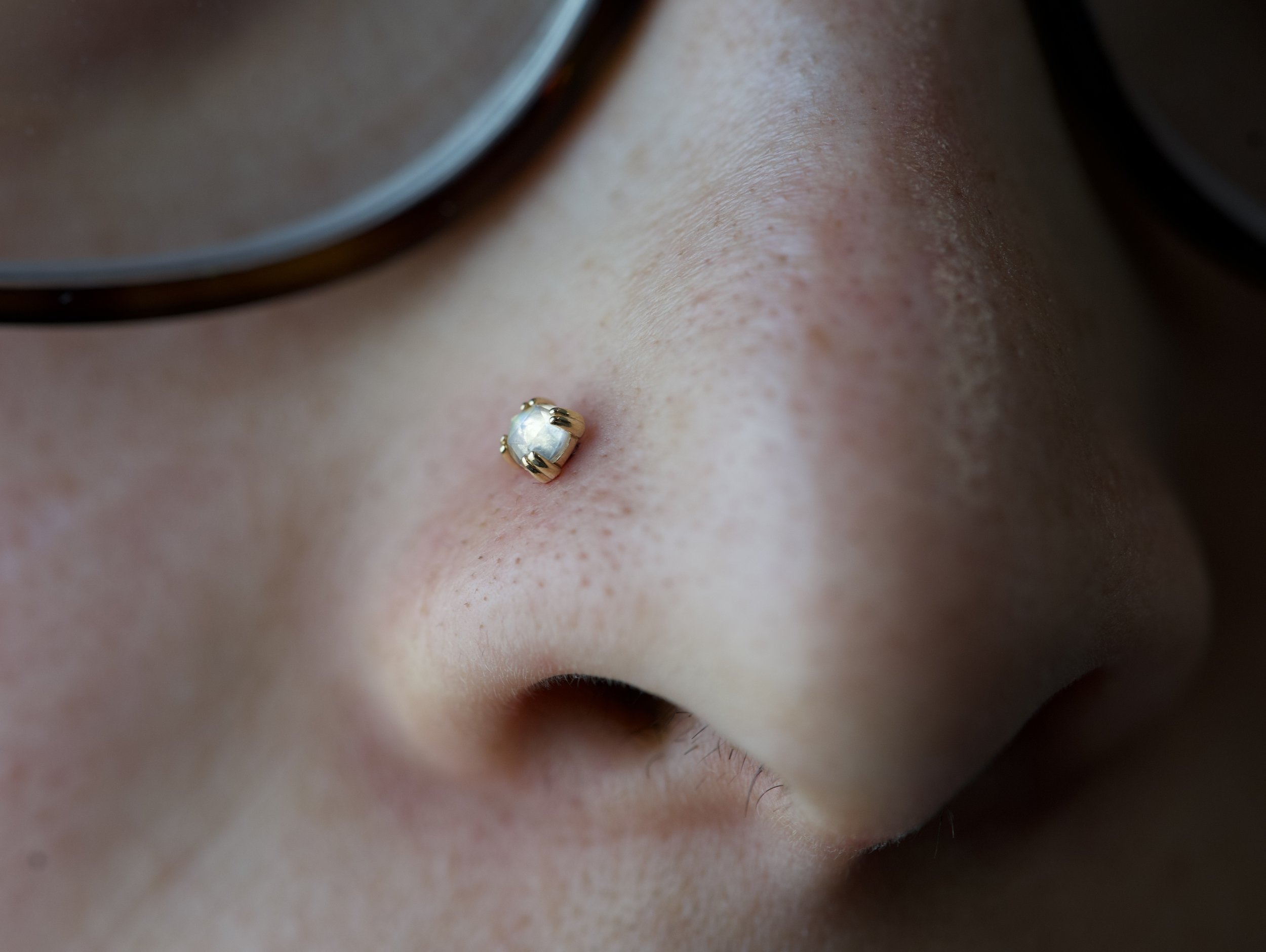Close-up of a person's nose with a gold nose stud featuring a clear gemstone, wearing glasses.