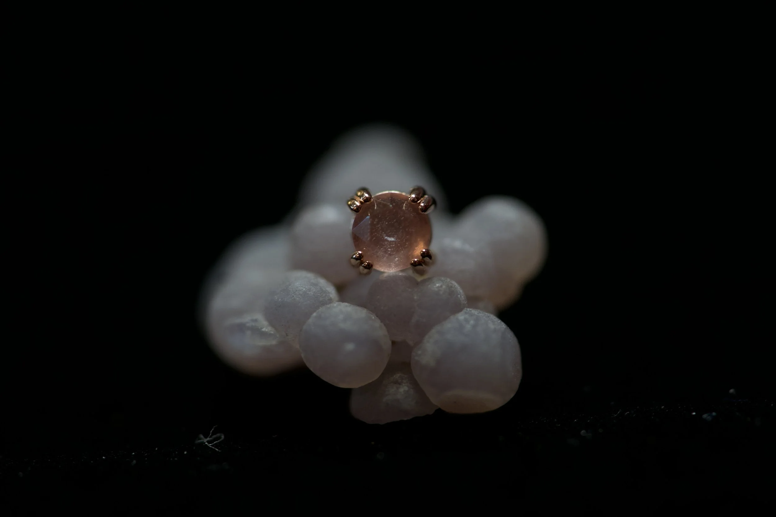 A close-up of a light pink gemstone set in a gold ring, resting on a white flower with multiple rounded petals against a dark background.