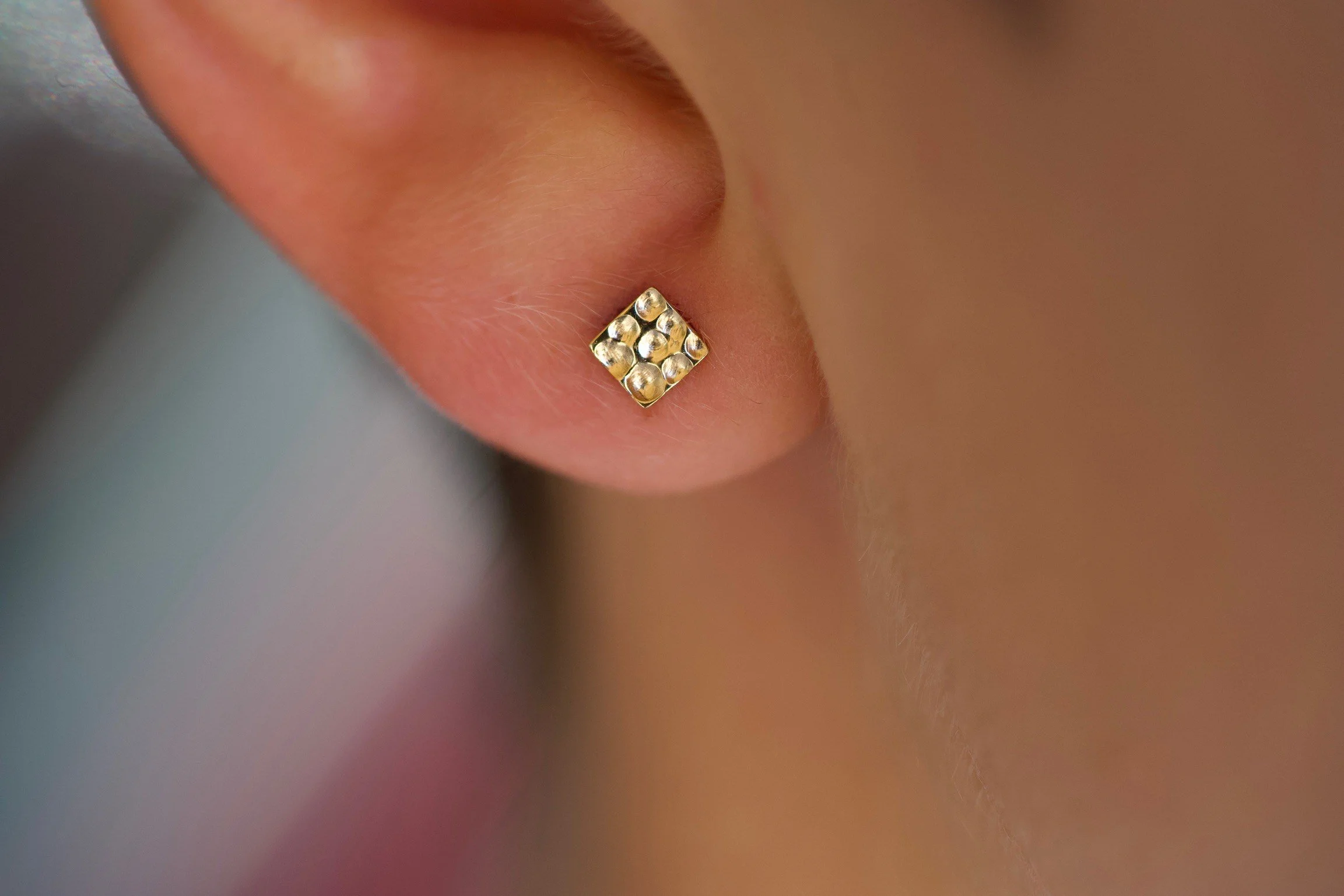Close-up of a person's earlobe with a gold stud earring featuring six small beads arranged in a square pattern.