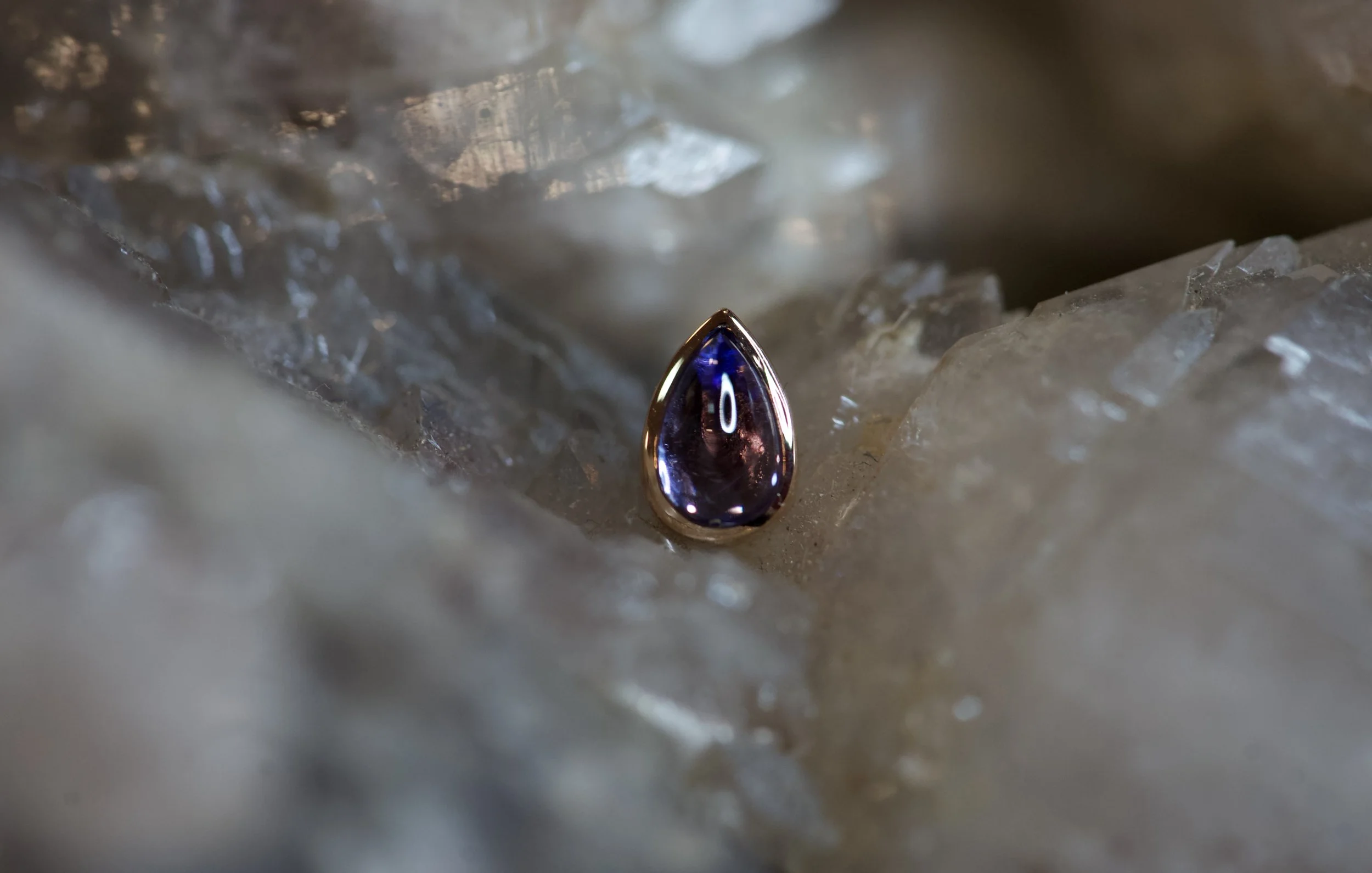 Close-up of a dark purple teardrop-shaped gemstone with a gold border, placed on clear ice crystals.