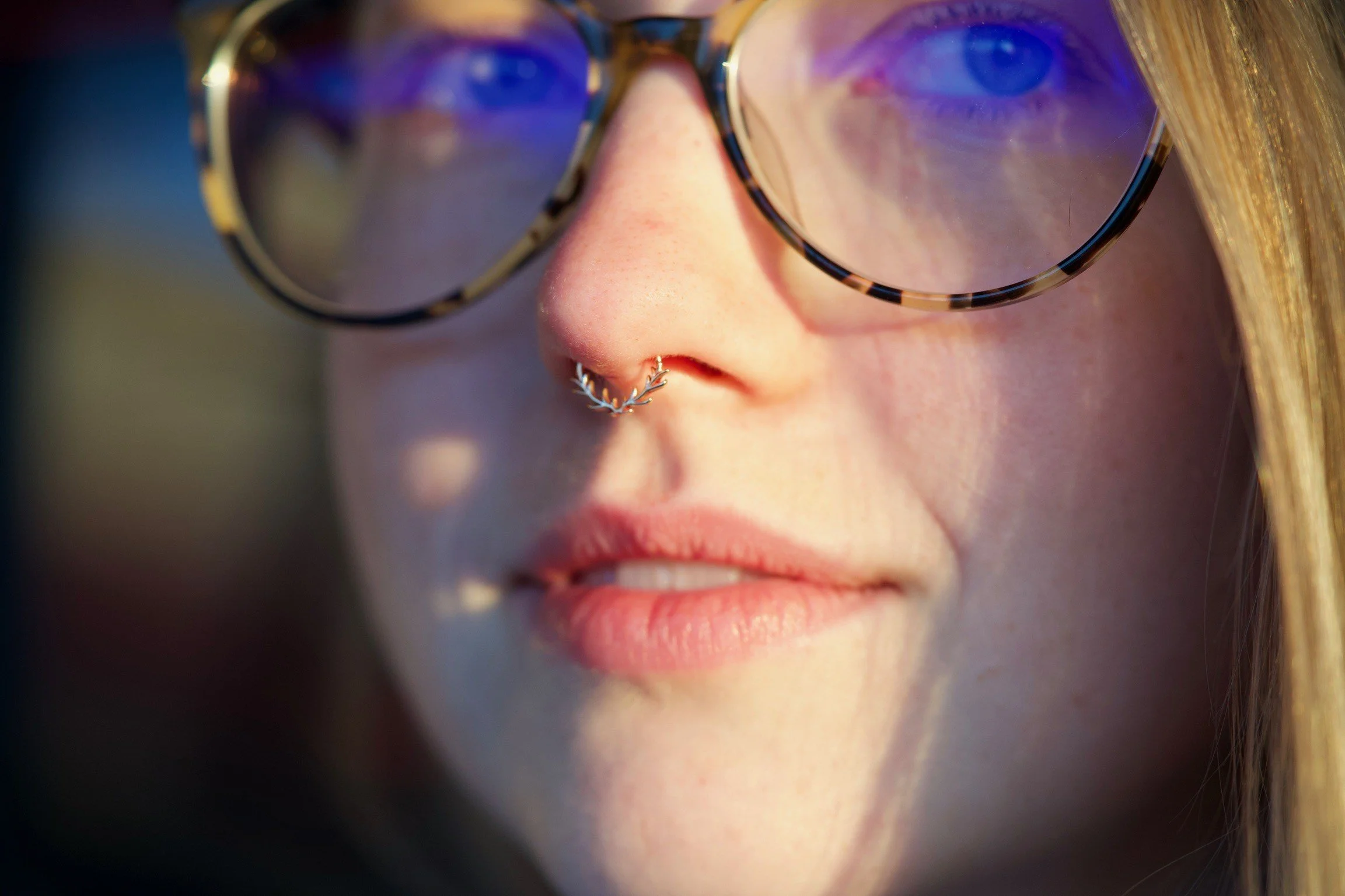 Close-up of a woman's face wearing large tortoiseshell glasses and a septum piercing shaped like antlers in bright, natural light.