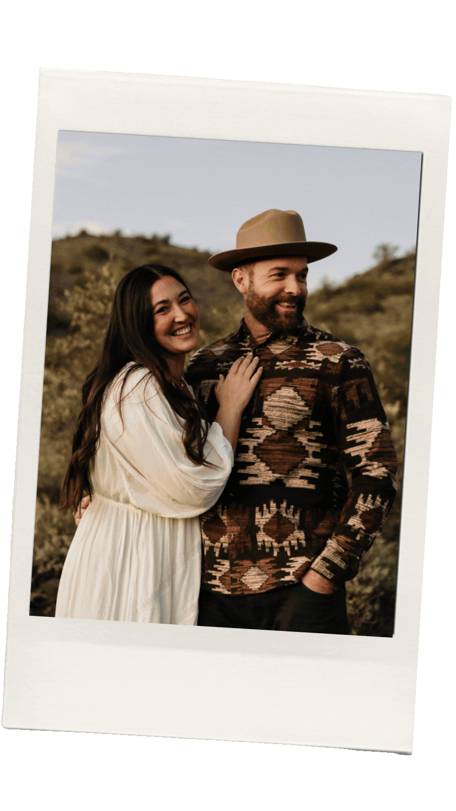A smiling woman with long dark hair and a man with a beard wearing a wide-brimmed hat and patterned shirt, standing outdoors with hills in the background.