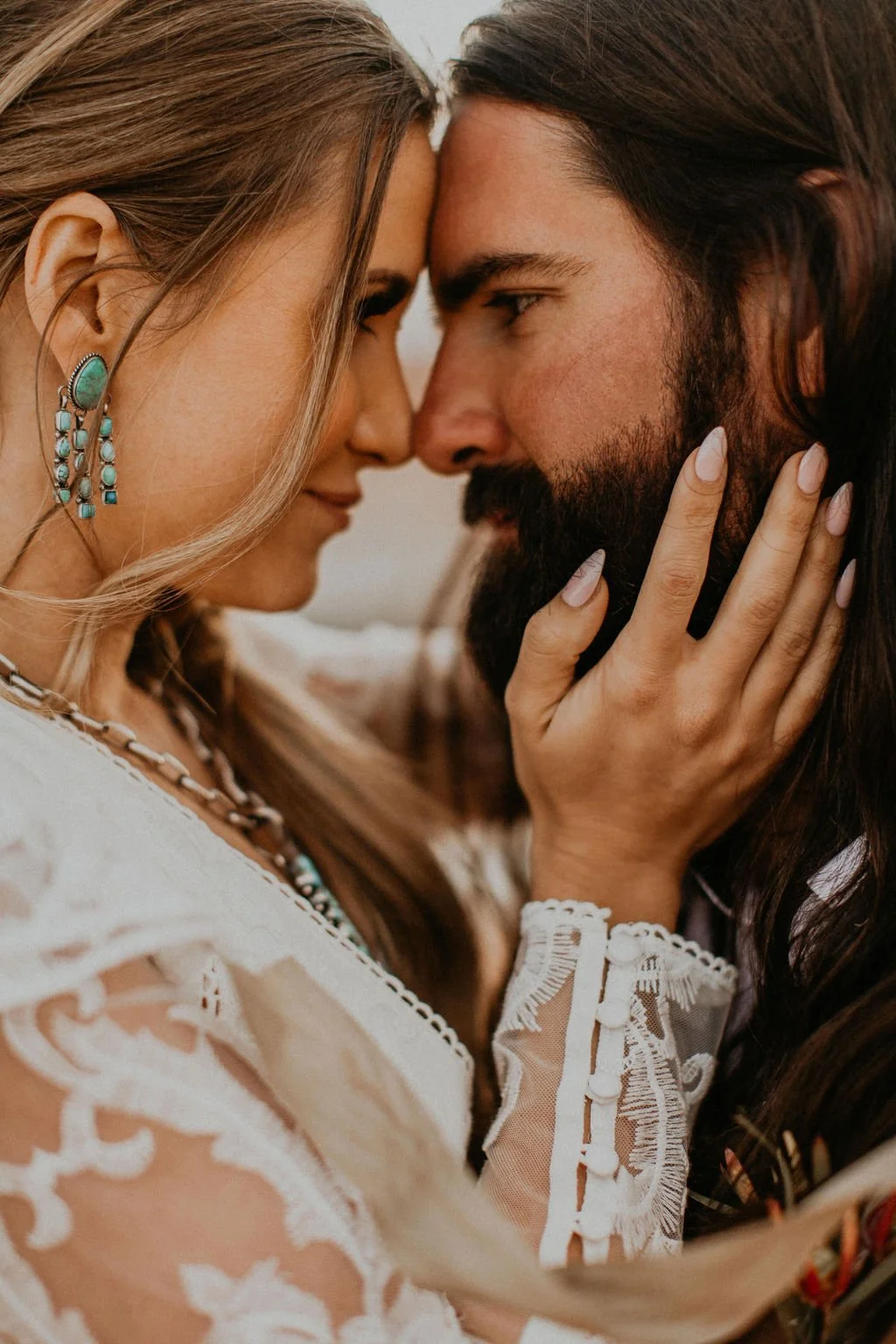 A close-up of a couple touching foreheads, the woman with blonde hair and turquoise earrings, and the man with long dark hair and a beard, holding each other's faces gently.