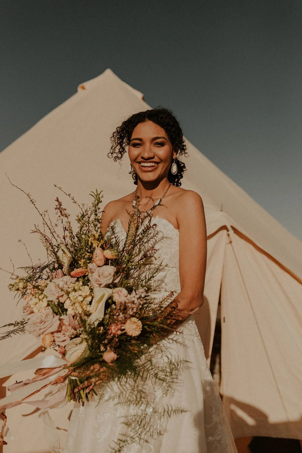 A woman in a wedding dress holding a bouquet of pink and white flowers, smiling in front of a beige tent under a clear blue sky.