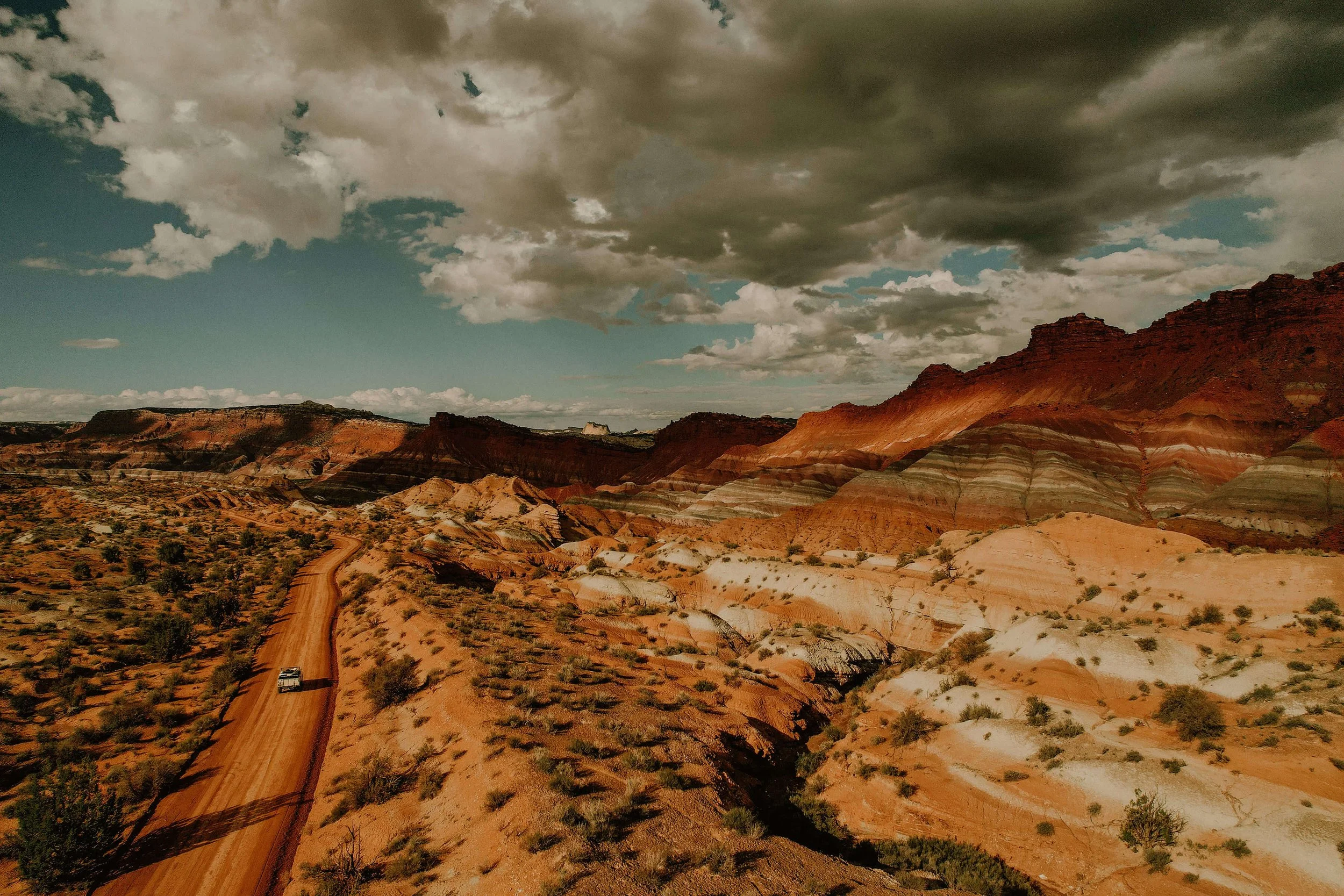A rugged desert landscape with multicolored rock formations, a dirt road winding through the terrain, and a dark, cloudy sky overhead.