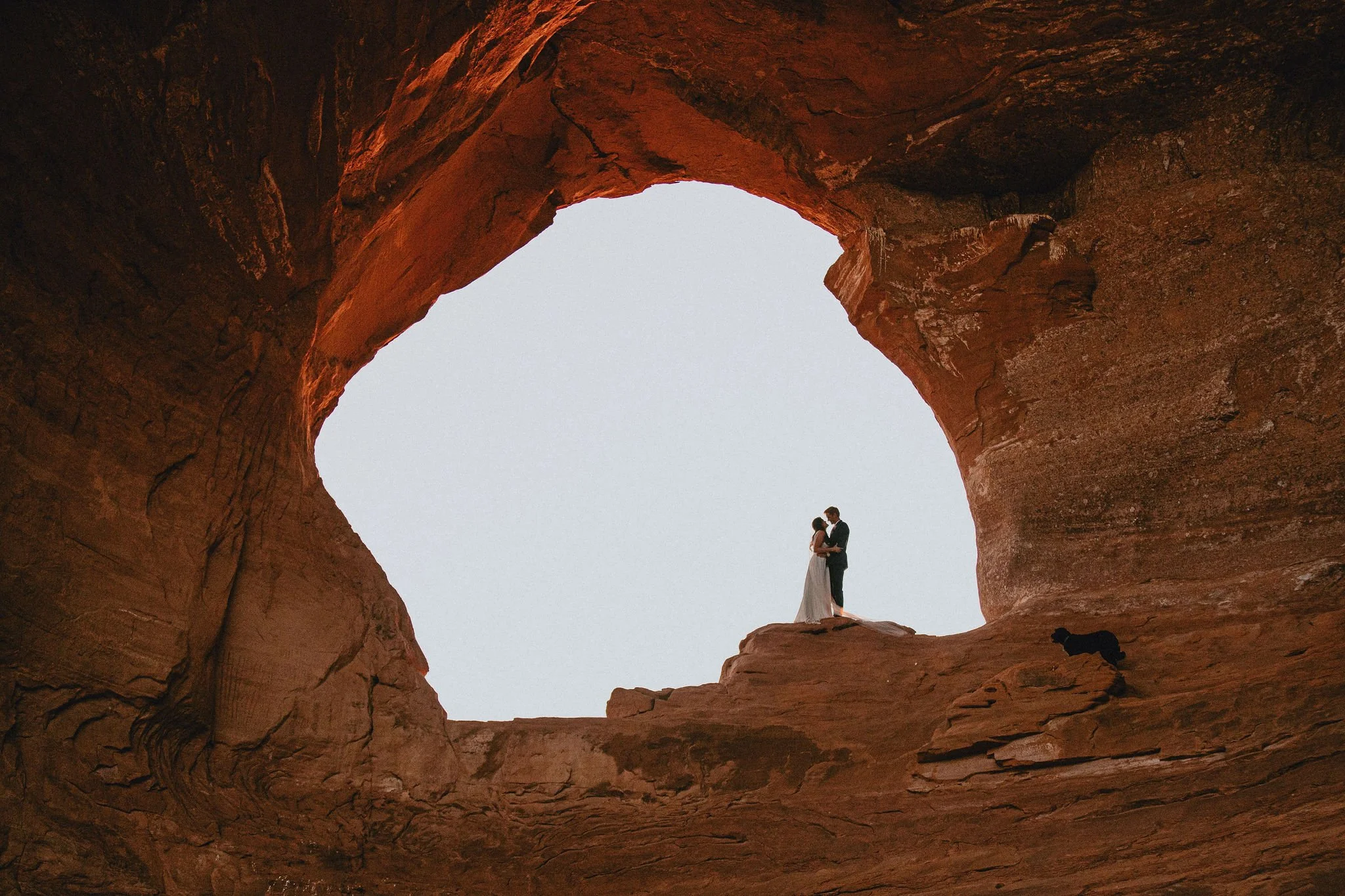 Couple in wedding attire standing inside a natural rock formation, with the sky visible through a large opening in the stone.