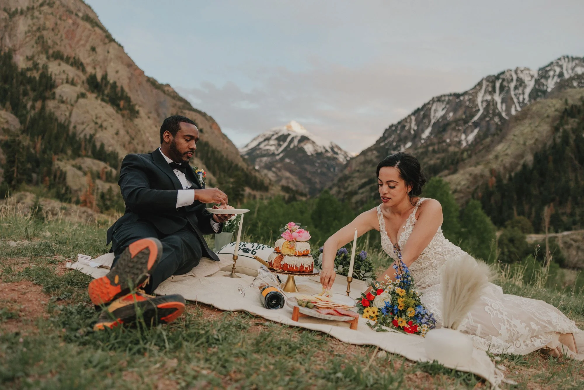 A man in a black tuxedo and a woman in a wedding dress having a picnic in the mountains in Colorado, with a cake, flowers, and food on a blanket.