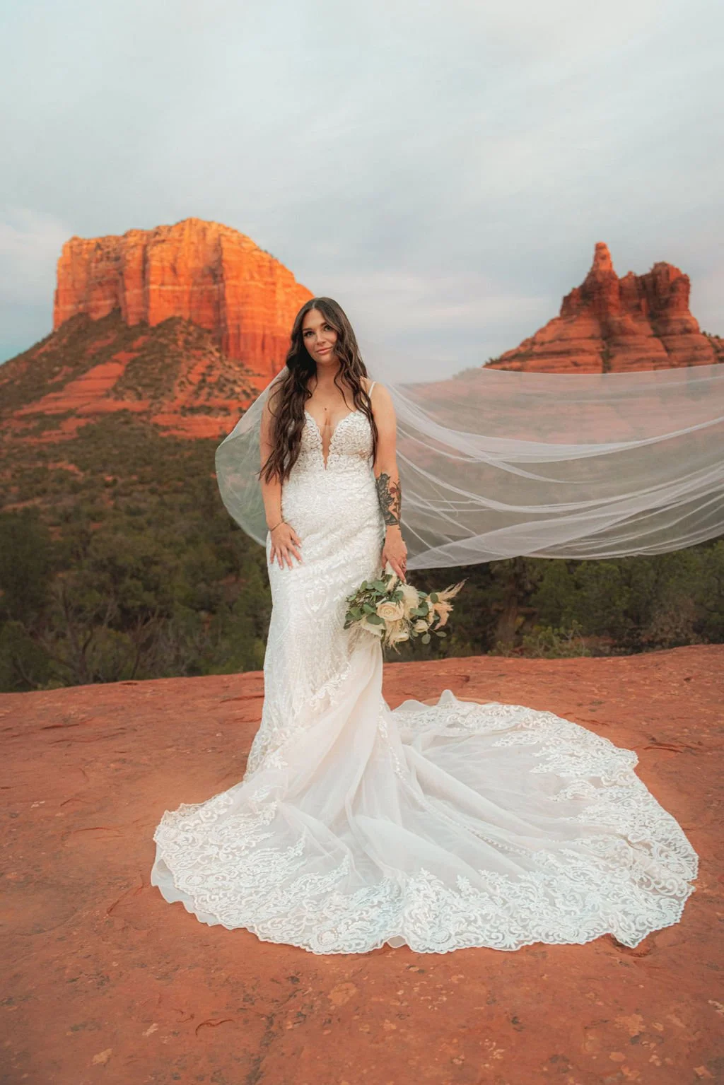 A bride in a white lace wedding gown holding a bouquet in a desert landscape with red rock formations in the background.