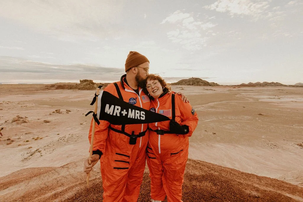 A couple dressed in orange space suits, standing on a desert-like landscape with rocky formations in the distance. They are embracing, and the man holds a flag that says "MR + MRS." He is kissing the woman's forehead, and they both are smiling.