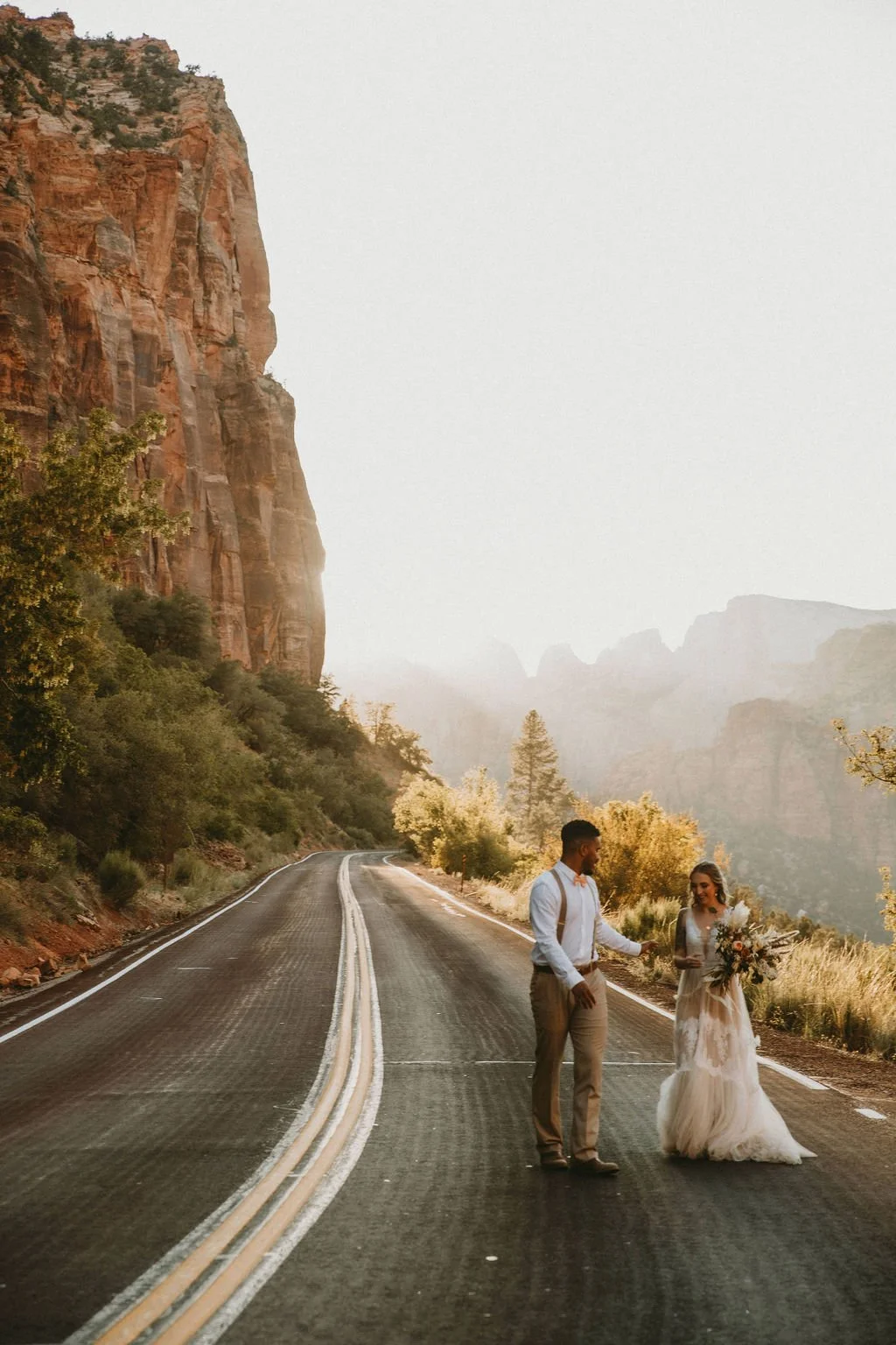 A couple in wedding attire standing on an empty, winding mountain road surrounded by tall cliffs and trees, with a hazy background of mountains and sky.