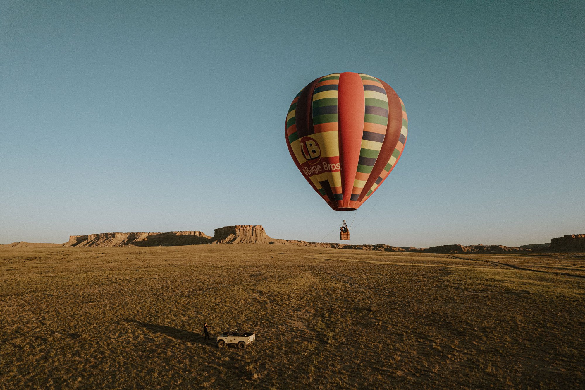 Hot air balloon with colorful stripes floating over a desert landscape with rock formations in the background during sunset.