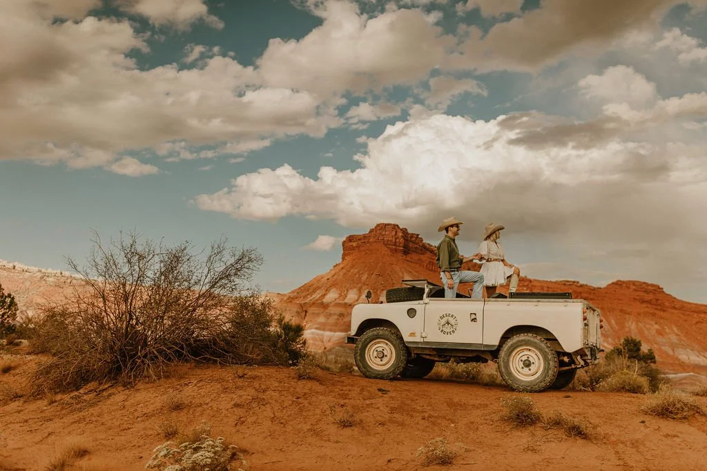 Two people in Western attire sit on the back of a white safari vehicle in a desert landscape with red cliffs and scattered bushes, under a partly cloudy sky.