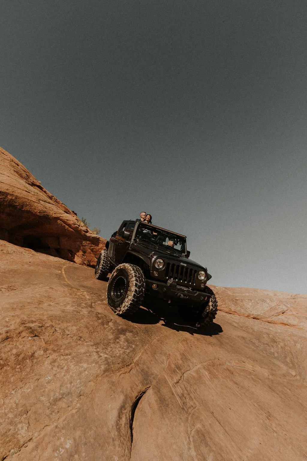 A black Jeep with large tires driving off-road on a rocky desert terrain, with two people inside, one of whom is standing up through the sunroof, taking a selfie or enjoying the view, under a clear blue sky.