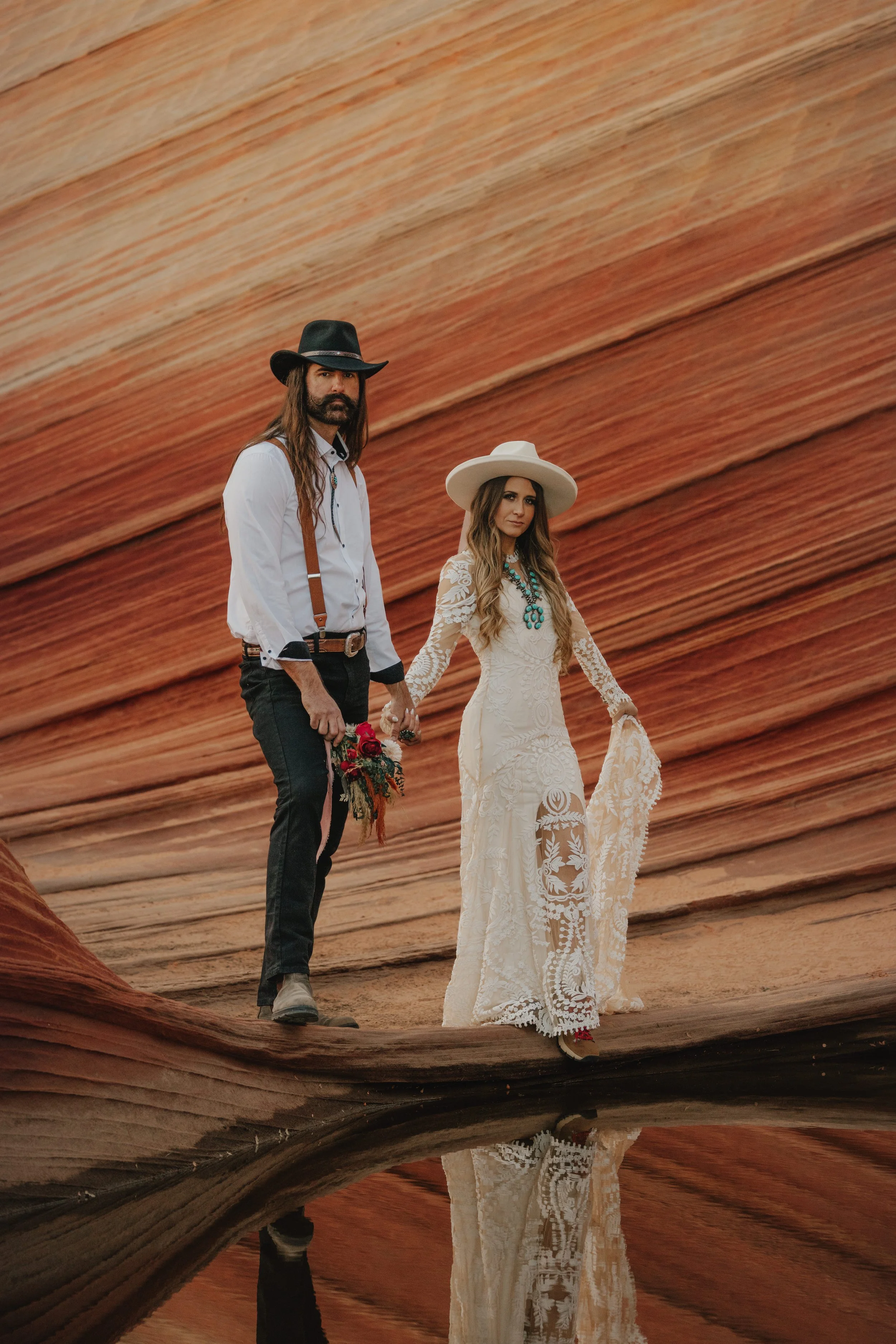 A bride and groom holding hands and smiling at each other in a desert landscape with rock formations and a cloudy sky.