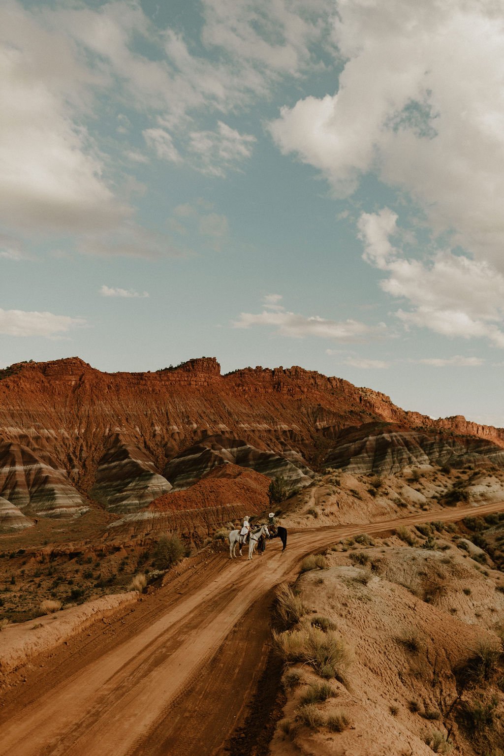 A dirt road curves through a desert landscape with rugged, multicolored hills and cliffs in the background. Two horseback riders with guides are on the road, and there are sparse desert shrubs and grasses. The sky is partly cloudy with a light blue h