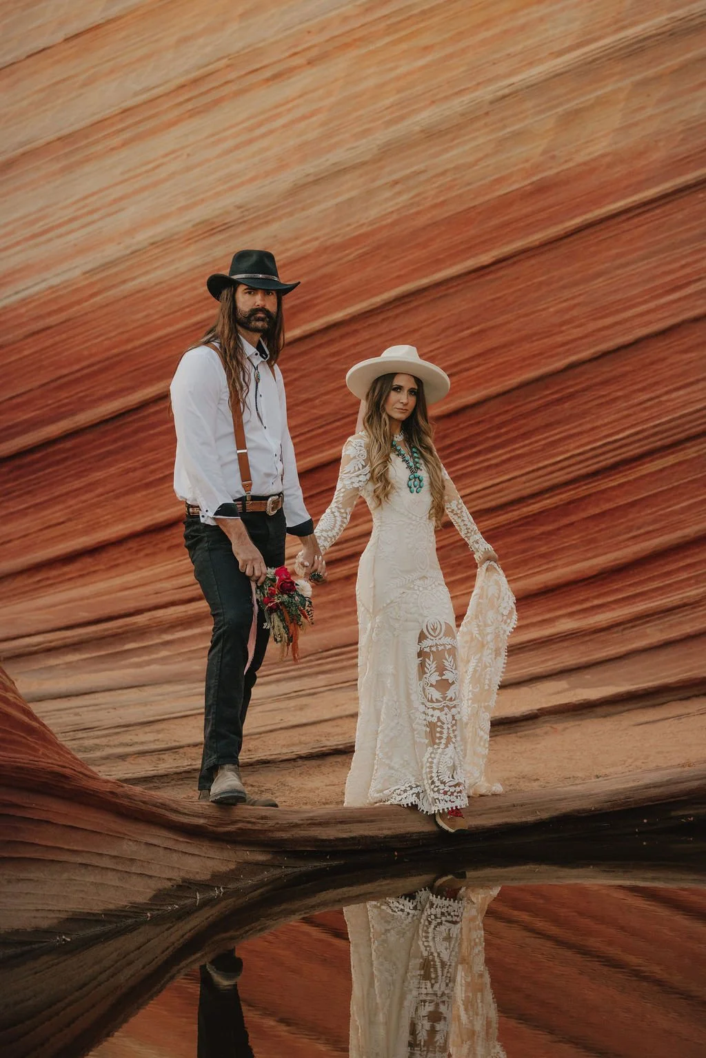 A man and woman hold hands in a desert landscape with rock formations. The woman wears a long white lace dress and wide-brimmed hat, and the man wears a cowboy hat, white shirt, black pants, and suspenders. They stand near a reflective water surface.
