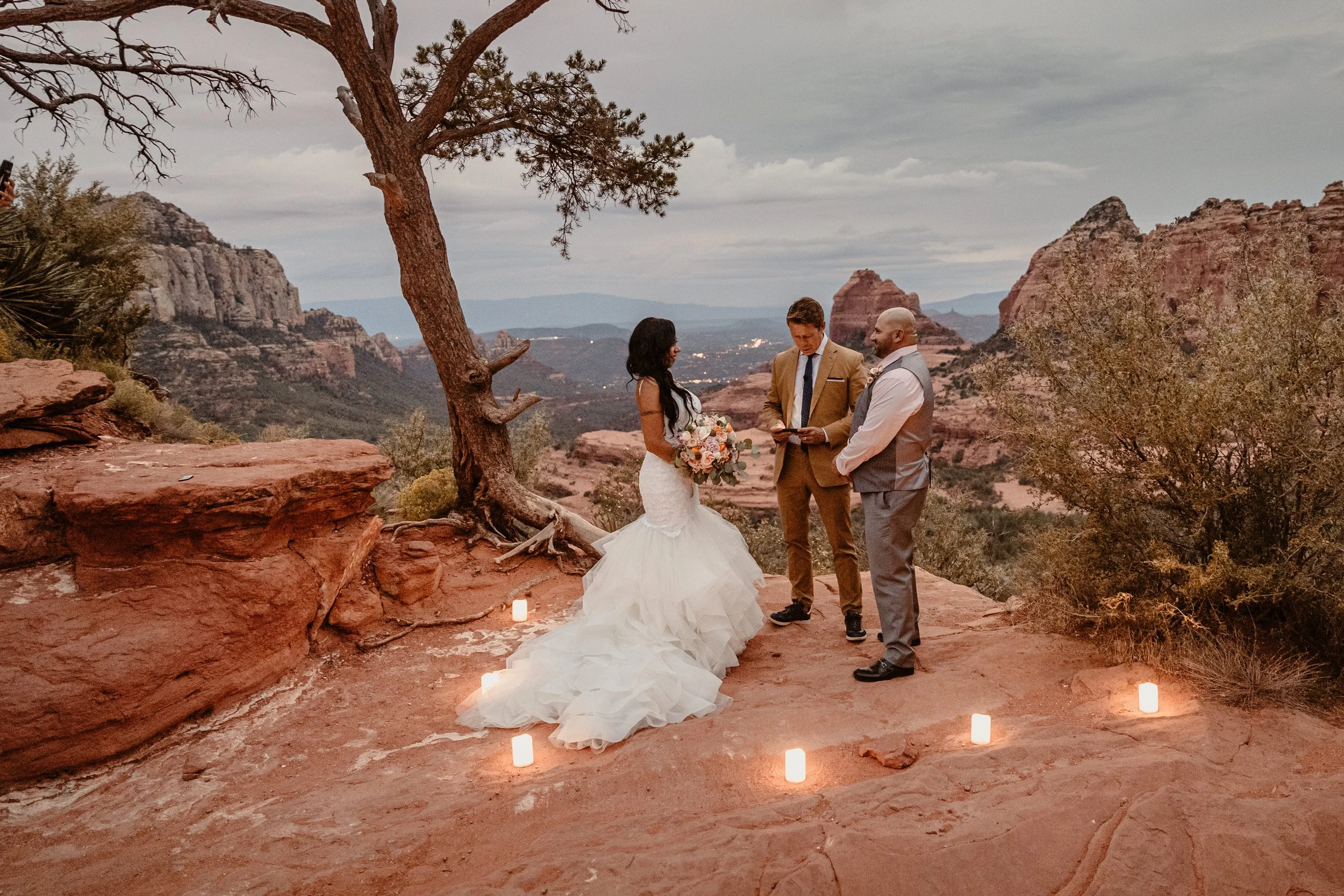 A wedding ceremony taking place outdoors on a rocky ledge with a mountainous desert landscape in the background. The bride in a white wedding gown holds a bouquet, and two grooms stand before her, one in a brown suit and the other in a gray vest. Can
