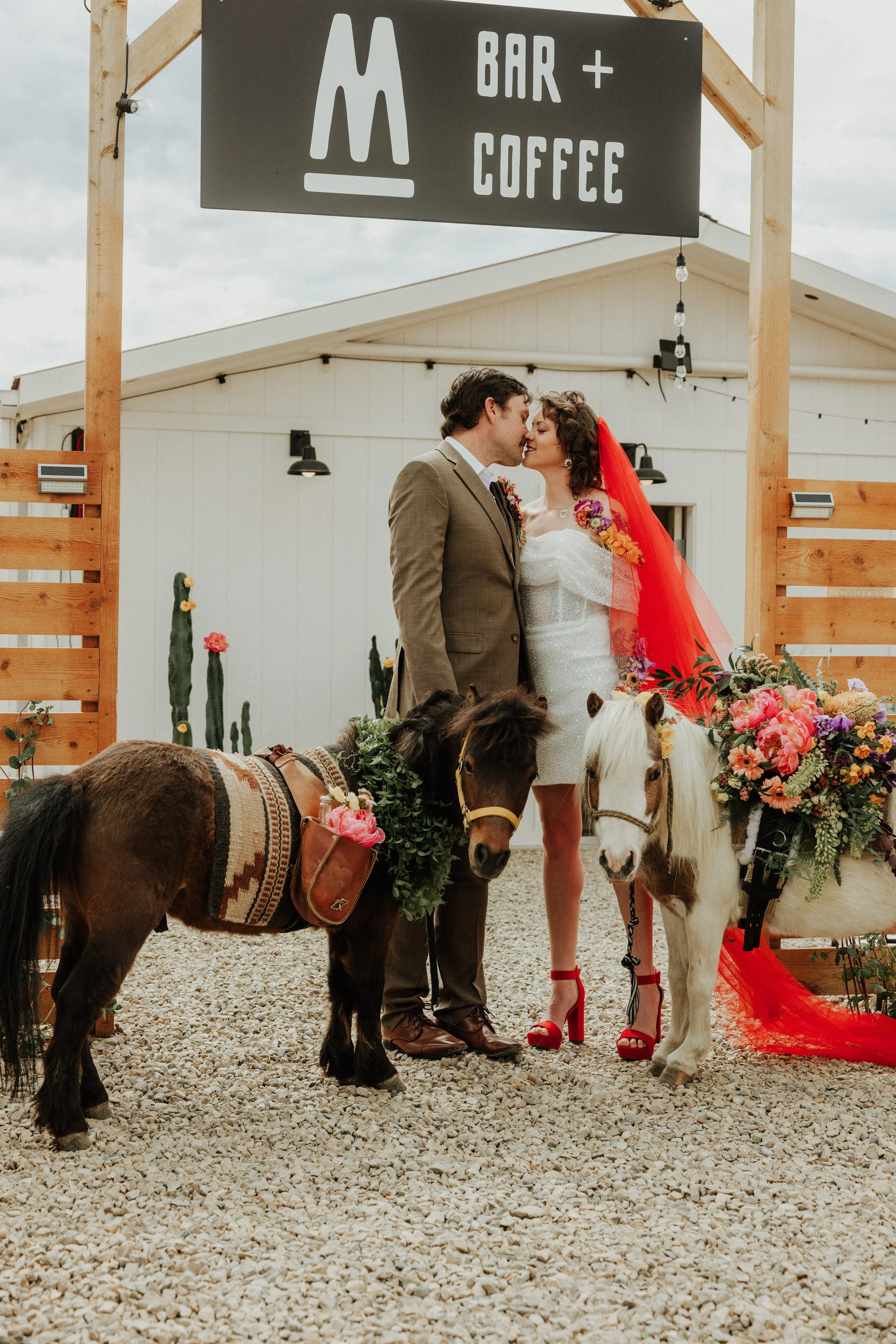 A couple in wedding attire sharing a kiss during their wedding ceremony, with two small ponies decorated with flowers in front of a wooden fence and a sign that reads 'Bar + Coffee.'