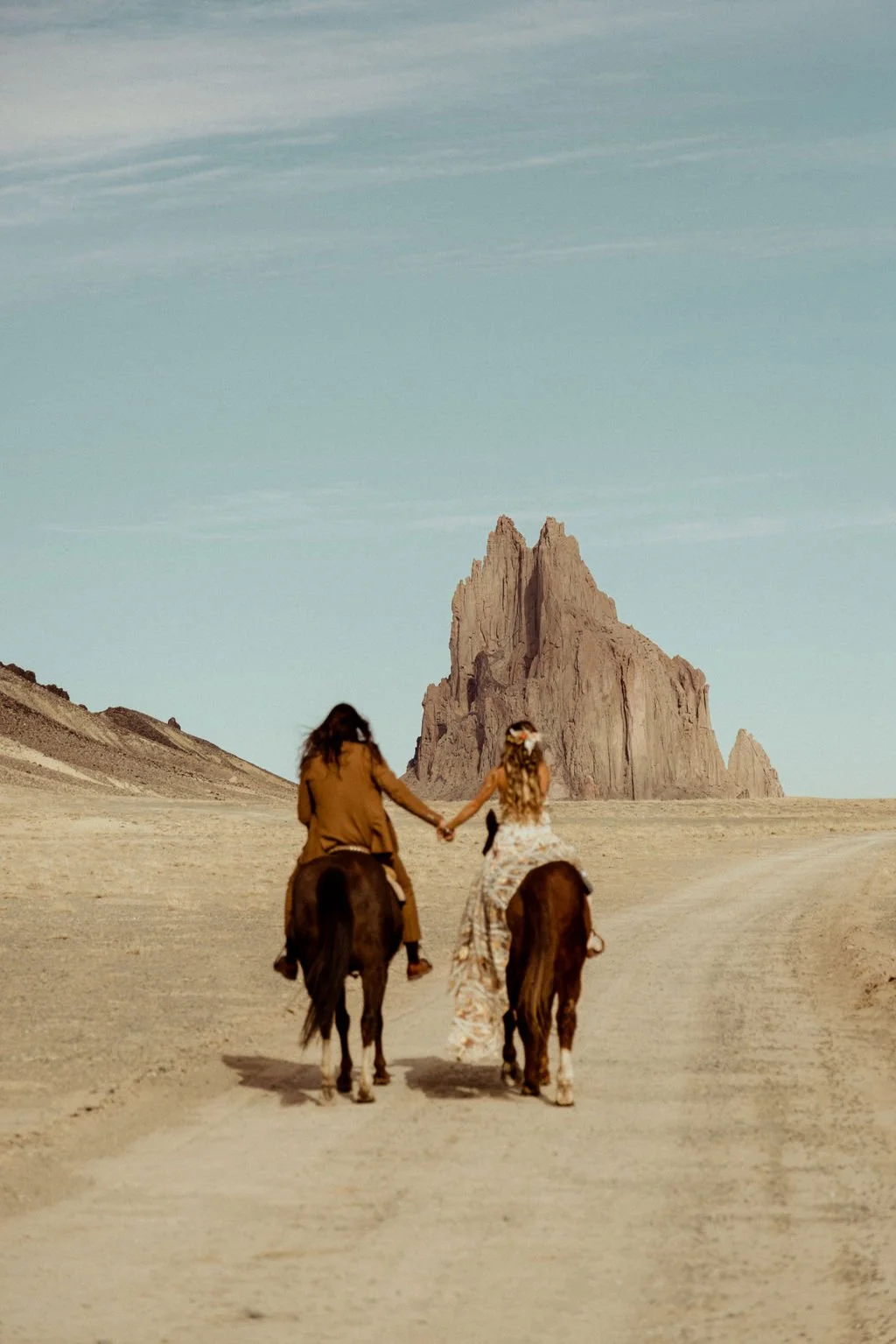Newlyweds riding horses in a desert landscape in New Mexico, holding hands, with a large rock formation in the background and a clear sky overhead.