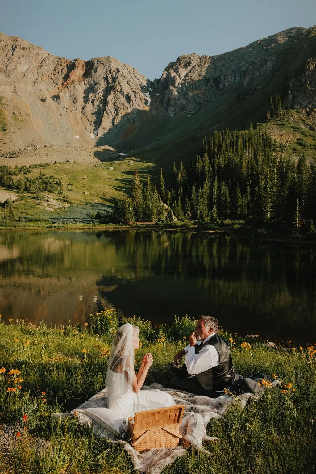 Couple having a picnic by a lake with mountains and evergreen trees in the background, during sunset.