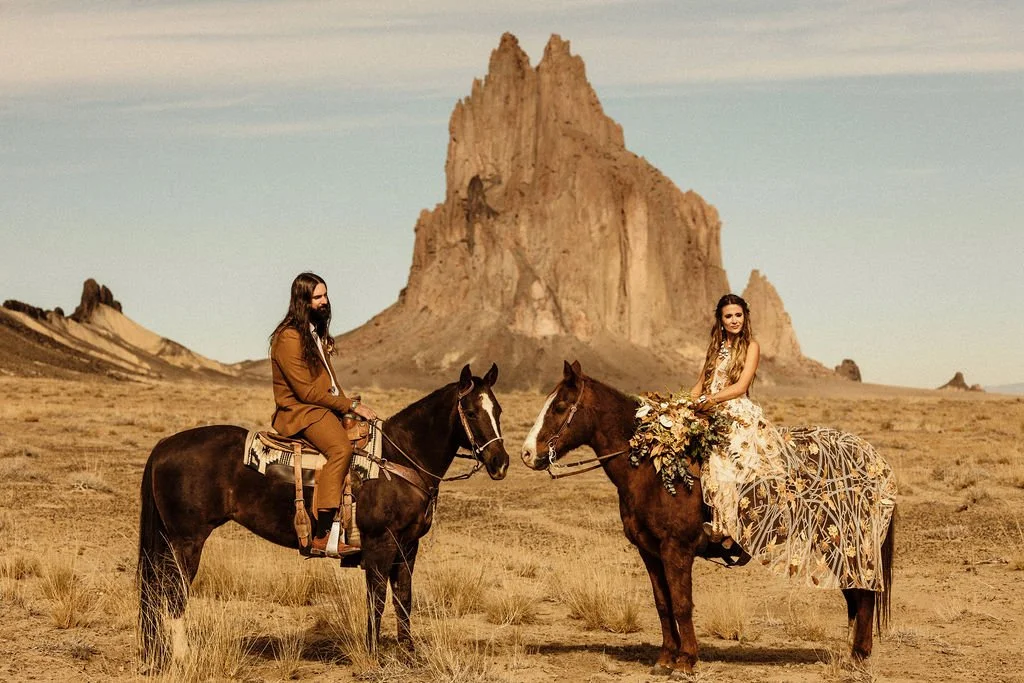 A man and woman riding horses in a desert landscape with a prominent mountain in the background. The woman is wearing a flowing dress and holding a large bouquet of flowers, while the man is dressed in a suit.