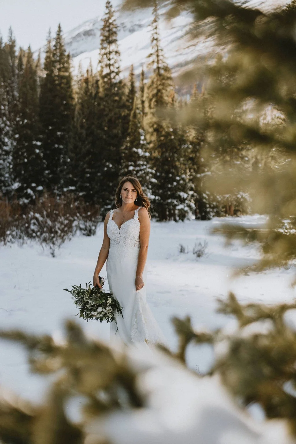 A bride standing in a snow-covered forest, wearing a white wedding gown and holding a bouquet of flowers.