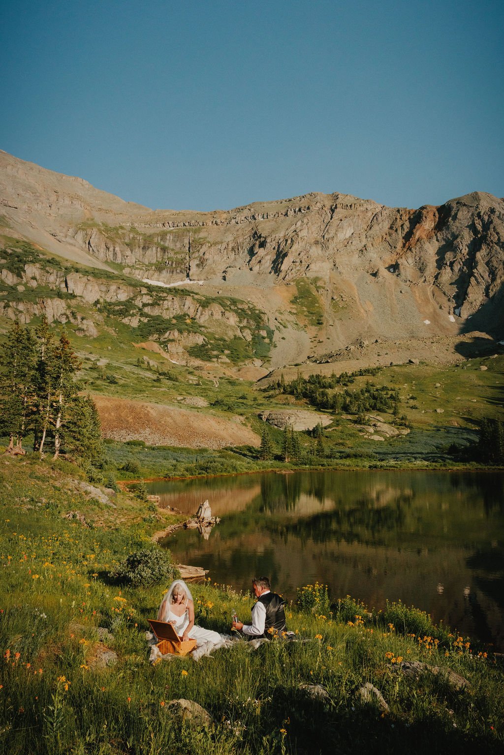 A couple having a picnic by a mountain lake surrounded by trees and green hills, with rocky mountains in the background on a clear day.