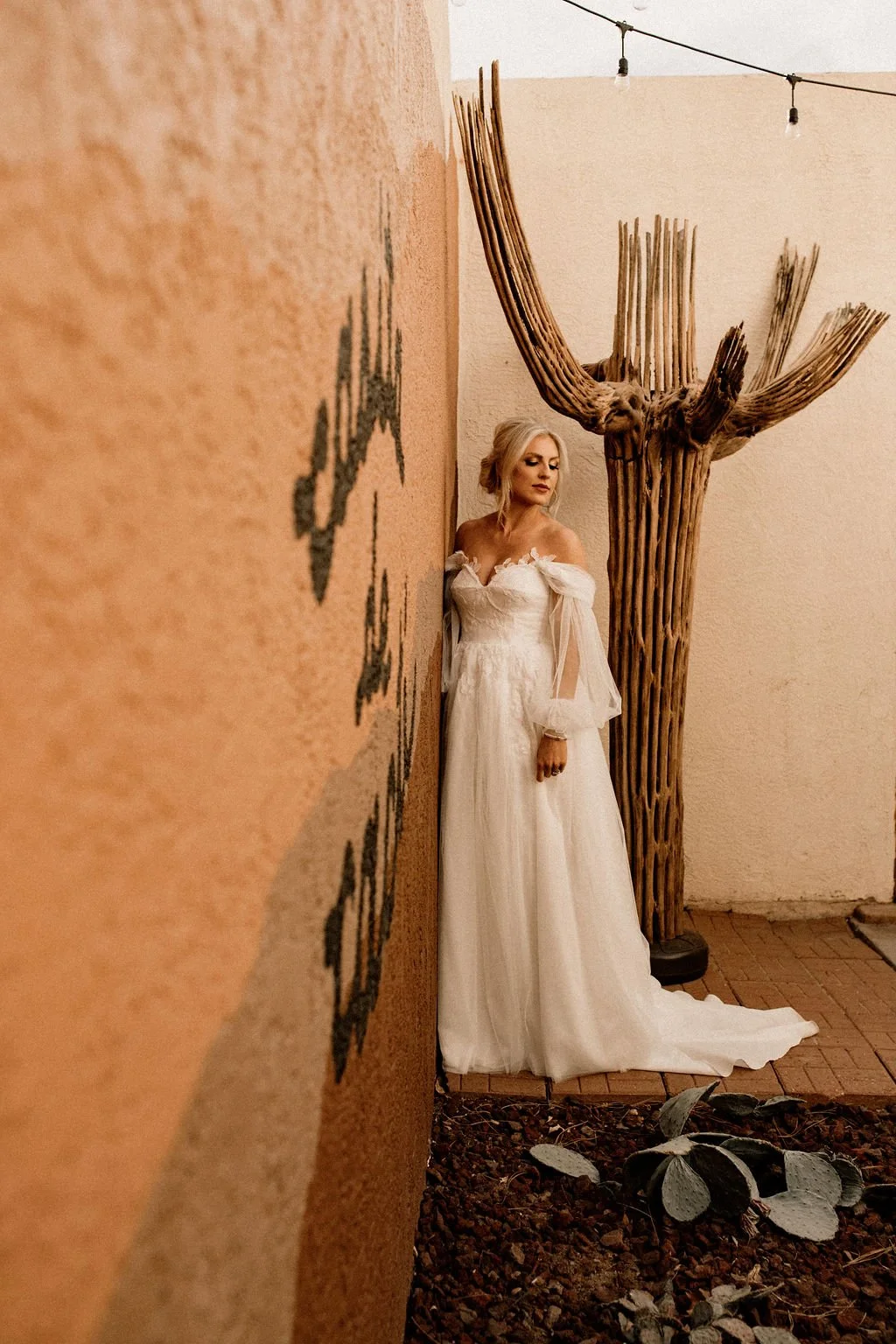 A bride in a white wedding dress leaning against a wall with a large wooden cactus sculpture in the background.
