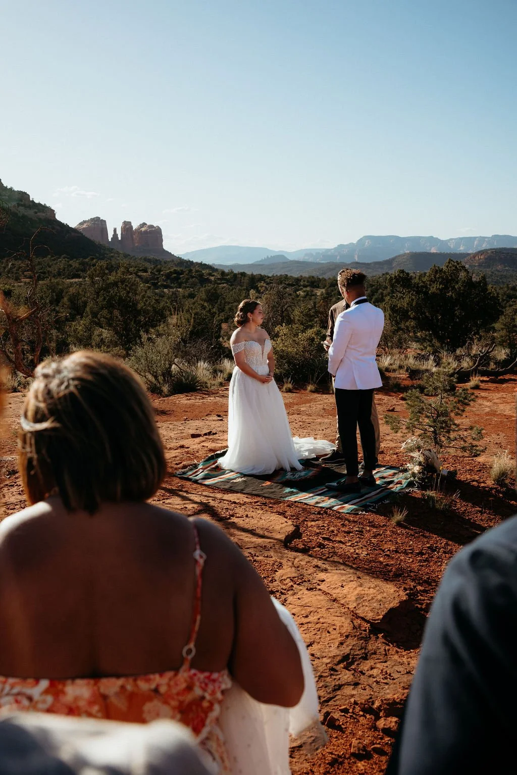 A wedding ceremony taking place outdoors on a rocky, desert landscape with mountains in the background. The bride and groom stand on a mat, exchanging vows, while guests sit and watch.